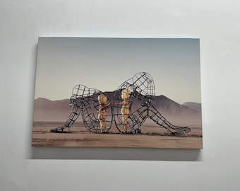 Op de afbeelding: Een zwart metalen sculptuur van drie figuren in een woestijnlandschap. De figuren zijn zo gepositioneerd dat ze elkaar omarmen of beschermen.