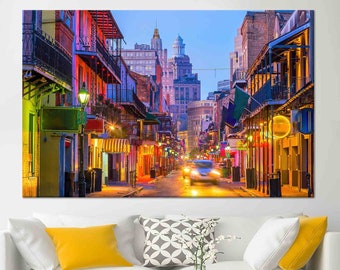 Puede incluir: Una escena callejera colorida en Nueva Orleans al atardecer. La calle está bordeada de edificios iluminados con balcones y barandillas de hierro forjado. Los edificios están pintados en una variedad de colores, incluyendo amarillo, rojo, verde y azul. La calle está vacía excepto por unos pocos coches en la distancia.