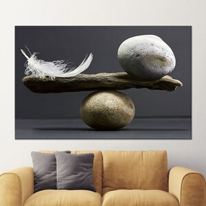May include: A still-life image features a balanced composition of natural elements. A white feather and a smooth, gray stone rest on a piece of driftwood, balanced atop a round, textured stone. The background is a solid, dark gray.