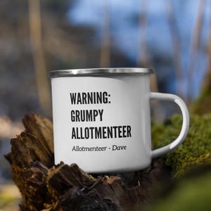 Może przedstawiać: Biały emaliowany kubek ze srebrnym rantem i uchwytem, ozdobiony czarnym napisem WARNING: GRUMPY ALLOTMENTEER oraz Allotmenteer - Dave. Kubek posiada gładkie wykończenie i klasyczny cylindryczny kształt przeznaczony do serwowania napojów.