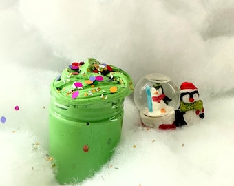 May include: Green slime with colourful sprinkles in a clear jar, a snow globe with a penguin inside, and a penguin figurine with a red hat on a white fluffy surface.