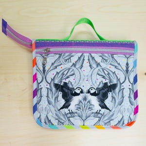 May include: A square, quilted pouch with a colorful, rainbow-striped edge and a green handle. The pouch features a zipper closure and a decorative pattern of two puffins facing each other, surrounded by leaves. The background is light gray.