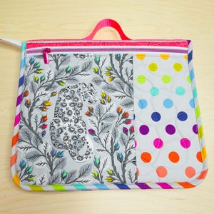 May include: A quilted tote bag with a colorful design. The bag features a leopard print panel, a polka dot panel, and a rainbow striped border. It has a pink zipper and a pink handle. The bag is made of fabric.