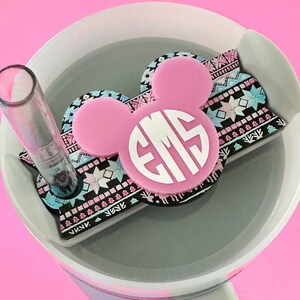 May include: A pink and white personalized Mickey Mouse-shaped tumbler topper with the initials "EMS" in white. The topper sits on a gray tumbler with a clear straw. The design includes a patterned border with blue, black, and pink accents.