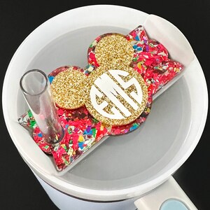 May include: A white and light blue travel mug with a decorative acrylic topper. The topper features a gold glitter circle, a white monogram, and a colorful confetti design. A clear straw is inserted into the mug.