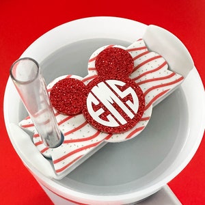May include: A white tumbler with a red and white striped Mickey Mouse-shaped lid and a clear straw. The lid features a red glitter monogram with the initials EMS. The tumbler sits on a red surface.