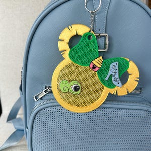 May include: A yellow and green circular keychain with a bow shape, featuring a small green chameleon, a cupcake, and a light blue high-heeled shoe with the text It's pos-si-ble! attached to a light blue backpack with a front pocket and silver zipper.