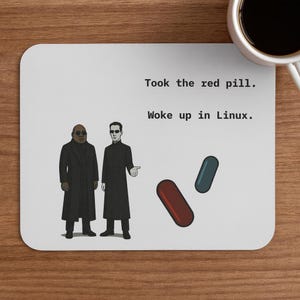 Könnte beinhalten: Weißes rechteckiges Mousepad mit einer Grafik von zwei Figuren in schwarzen Mänteln und Sonnenbrillen, mit roten und blauen Pillen. Der Text lautet: "Took the red pill. Woke up in Linux."