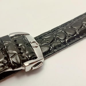 May include: Close-up of a black watch strap with a crocodile skin texture. The strap features a silver-colored buckle. The watch band is angled across the frame, showcasing the details of the leather and the metal clasp.