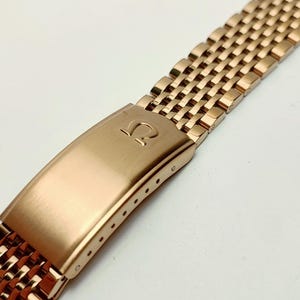 Pode incluir: Uma pulseira de relógio em ouro rosa com um design de malha. A pulseira apresenta um fecho retangular com o logotipo da Omega. A pulseira é feita de pequenas ligações interconectadas, criando uma superfície flexível e texturizada.