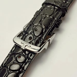 Peut inclure: Gros plan sur un bracelet de montre noir avec un motif texturé en forme de peau de crocodile. Le bracelet est doté d'une boucle argentée au design distinctif. Le bracelet de montre est incliné sur un fond blanc.
