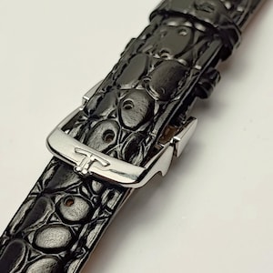 Könnte beinhalten: Ein schwarzes Uhrenarmband mit einer strukturierten, geprägten Krokodiloptik und einer polierten silberfarbenen Metallschnalle. Die Schnalle zeigt ein stilisiertes T-förmiges Logo auf der Vorderseite und sichert das Band mit einem Dorn.