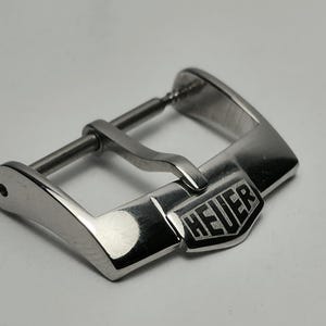 May include: A silver-colored watch buckle with the brand name "HEUER" engraved on the front. The buckle has a rectangular shape with rounded edges and a bar for the watch strap.