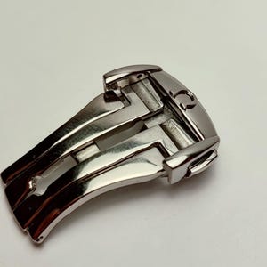 Può includere: Una chiusura per orologio argentata, con un design curvo e un meccanismo a cerniera. La chiusura ha una finitura lucida e un piccolo dettaglio inciso. La chiusura è progettata per fissare un cinturino.