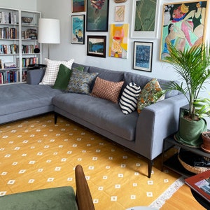 Op de afbeelding: Een grijze hoekbank met sierkussens op een gele vloerkleed met witte ruitpatronen. Verschillende ingelijste kunstwerken sieren de muur. Een potplant en een bijzettafel bevinden zich rechts van de bank.