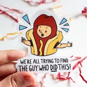 May include: A sticker featuring a cartoon hot dog character with a surprised expression, dressed in a yellow bun and red hat. The text on the sticker reads, "WE'RE ALL TRYING TO FIND THE GUY WHO DID THIS!" The sticker is held by a hand.