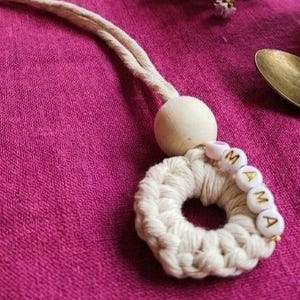 Peut inclure: Un porte-clés macramé blanc fait à la main avec le mot "MAMA" en perles dorées. Il comporte une perle en bois et une gland. Posé sur un tissu magenta, à côté d'une tasse en céramique et d'une cuillère.