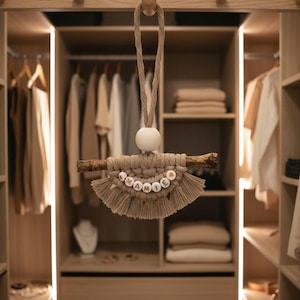 Peut inclure: Suspension murale en macramé beige avec le mot "MAMIE" en perles de bois. Le macramé est fixé à une branche en bois naturel et suspendu par une corde beige et une perle en bois blanche. En arrière-plan, une garde-robe en bois.