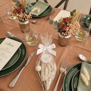 Peut inclure: Une table dressée avec des assiettes vertes, des couverts et des verres. Deux petits arrangements floraux dans des vases en verre mercurisé sont sur la table. Un petit sac cadeau avec un motif macramé et les lettres "COJ" est au centre.