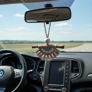 Peut inclure: Un charm de voiture en macramé beige suspendu au rétroviseur d'une voiture. Le charm comprend une baguette en bois, une perle blanche et un design en forme d'éventail avec le mot "dream" épelé en petites perles rondes. Le tableau de bord et le volant de la voiture sont visibles.
