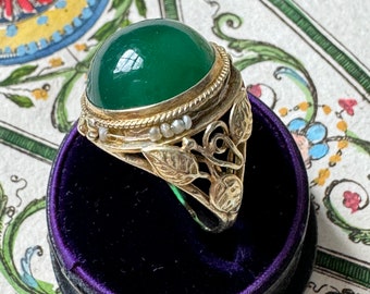 Pode incluir: Um anel de ouro com uma gema verde e detalhes de pérolas. O anel é engastado em um design de folhas e é exibido em uma caixa de anel de veludo roxo.