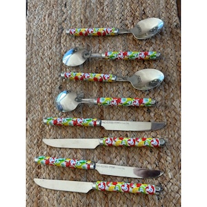 May include: A set of stainless steel spoons and knives with colorful handles. The handles feature a repeating pattern of red, yellow, and green fruit and faces. The knives have the text "FAC-022583-17295" printed on the blade.