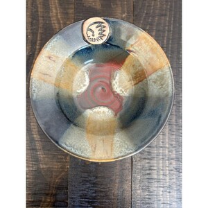 May include: A handmade ceramic bowl with a unique design. The bowl features a circular shape with a central red spiral and sections of blue, tan, and brown. A small, round emblem with a Native American design and the year 2019 is attached.