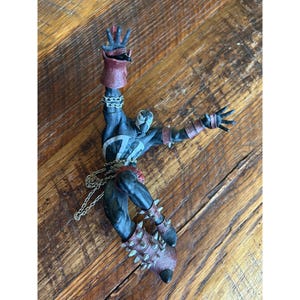 May include: A Spawn action figure in a dynamic pose, featuring a black and blue suit with a white emblem, red accents, and spiked boots. The figure is detailed with chains and a white mask, set against a wooden surface.