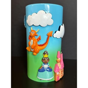 Op de afbeelding: Een cilindrische, blauwe keramische container met een grillig ontwerp. Het toont een cartoon draak, prinses, kasteel en wolken. De draak is oranje, de prinses draagt een kroon en blauwe jurk, en het kasteel is roze.