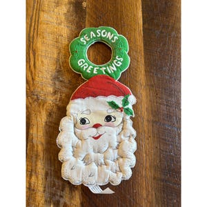May include: A vintage Santa Claus Christmas decoration. The Santa face is white with a red hat and a green holly accent. The top has a green ring with the words "SEASONS GREETINGS" in white.