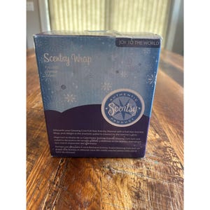 May include: A rectangular box for a Scentsy Wrap, with the text "Scentsy Wrap" and "Joy to the World". The box is blue with snowflake designs and an "Authentic Scentsy Product" seal. The box is on a wooden surface.