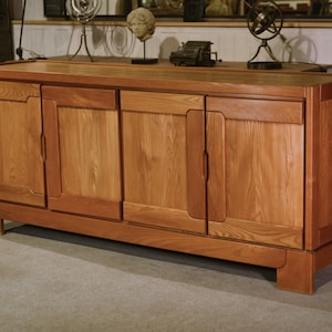 Peut inclure: Un buffet en bois brun clair à quatre portes. Le meuble est de forme rectangulaire et est fait de bois avec une finition naturelle. Les portes ont un grain de bois vertical et des panneaux encastrés. Style milieu du siècle.