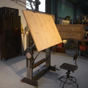 May include: An antique drafting table with a large, light-coloured wooden surface and adjustable metal frame. A matching dark wooden chair sits nearby. The table features a mechanical arm and a roll holder. The table is labelled "UNIC".
