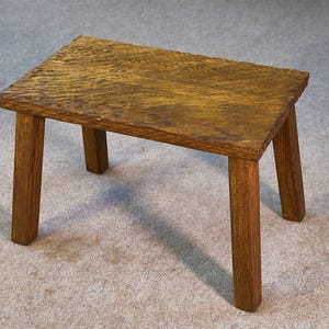 Peut inclure: Un petit tabouret en bois avec un dessus rectangulaire et quatre pieds robustes. Le bois présente une teinte brune chaude avec un grain et une texture visibles. Le tabouret est conçu pour un usage fonctionnel et ajoute une touche rustique.