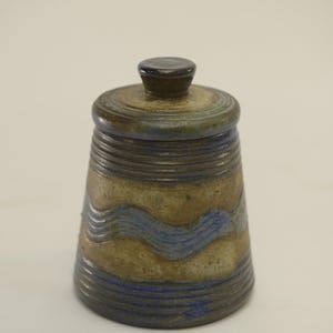 Peut inclure: Un petit pot en céramique avec un couvercle. Le pot est brun avec des lignes ondulées bleues et blanches et un bord bleu. Le couvercle est brun avec un dessus bleu.