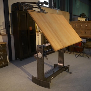 Peut inclure: Une table à dessin vintage avec un plateau en bois clair et une base en métal. La table est inclinée vers le haut, mettant en valeur sa conception réglable. La base en métal a une finition sombre et vieillie, dans une pièce bien éclairée.