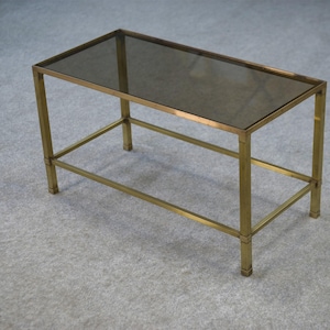 May include: A rectangular coffee table with a smoked glass top and a brass-colored metal frame. The table has a simple, minimalist design with straight lines and a lower support bar. The table measures approximately 36 inches long, 18 inches wide, and 16 inches high.