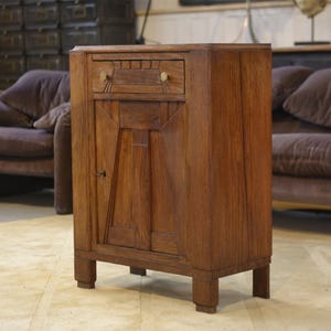 Puede incluir: Un mueble de madera vintage de color marrón cálido, con un cajón y una puerta. El mueble tiene un diseño geométrico en la puerta y se apoya en cuatro patas cortas. El cajón tiene un pequeño pomo redondo.