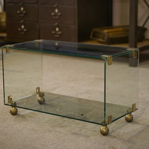 Peut inclure: Table basse rectangulaire en verre avec des coins et des pieds sphériques en laiton. Le verre transparent permet de voir l'intérieur. La table est sur roulettes, et le design général est minimaliste et moderne.