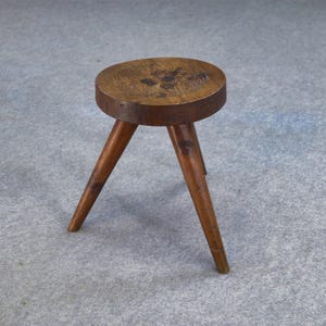 Peut inclure: Petit tabouret en bois à trois pieds avec une assise ronde. Le bois clair présente des veines et des nœuds. Les pieds sont inclinés vers l'extérieur. Un meuble rustique et artisanal, idéal pour divers usages.