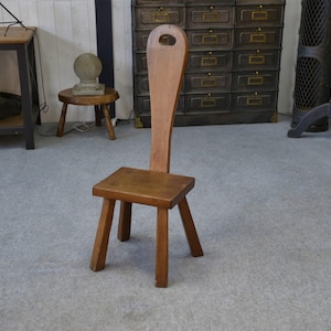 May include: A small wooden chair with a tall back and a square seat. The chair has three legs and a handle-shaped cutout at the top of the backrest. The wood has a warm, brown tone.