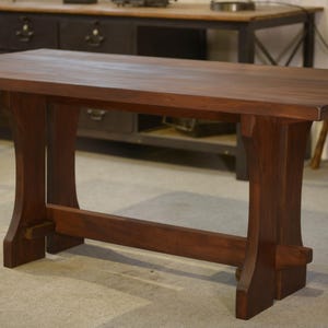 Peut inclure: Table en bois brun foncé avec un plateau rectangulaire et un piètement en chevalet. La table a une surface lisse et des pieds inclinés, reliés par une poutre horizontale. La table mesure environ 183 cm de long.