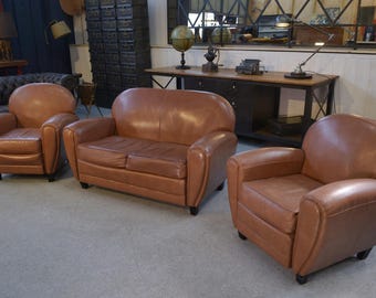Puede incluir: Un conjunto de tres muebles tapizados en cuero marrón. El conjunto incluye dos sillones y un sofá de dos plazas. Los muebles tienen un diseño redondeado y patas de madera oscura. Las piezas están dispuestas en una habitación con suelo de color neutro.