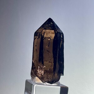 Può includere: Una punta di cristallo di quarzo fumé marrone scuro traslucido con una terminazione sfaccettata appuntita. Il cristallo presenta inclusioni interne naturali e striature verticali lungo i lati, montato verticalmente su una base quadrata in vetro.