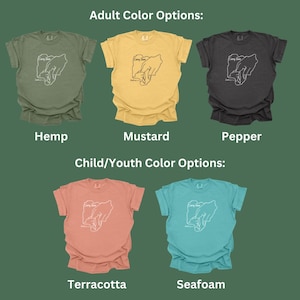 May include: A graphic t-shirt design featuring a line drawing of two hands holding each other. The design is available in a variety of colours for adults and children. The adult colours are hemp, mustard, and pepper. The children's colours are terracotta and seafoam.