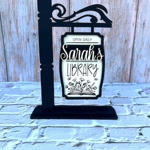 May include: Black wooden sign with the text "Sarah's Library" in a decorative font. The sign features a floral design and the words "Open Daily". The sign is mounted on a black post with a decorative top.
