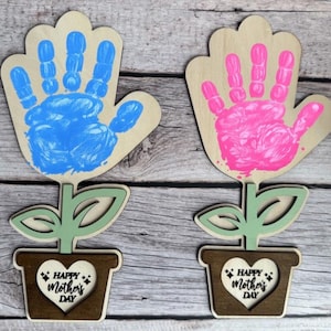 May include: Two wooden flower-shaped decorations with handprint designs. One flower has a blue handprint, the other a pink handprint. Each flower sits in a brown pot with a heart-shaped cutout that reads "Happy Mother's Day."