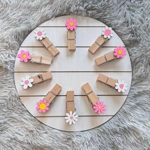 May include: A set of 10 wooden clothespins with pink and white flower embellishments. Each clothespin has a different flower design, including a heart in the center of one flower.