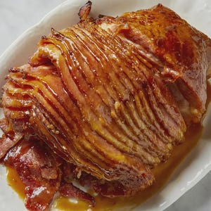 Puede incluir: Un jamón glaseado, cortado en rodajas y dispuesto en una fuente blanca con borde festoneado. El jamón tiene un color marrón dorado intenso con capas visibles y un glaseado brillante. La fuente está sobre una superficie de mármol.