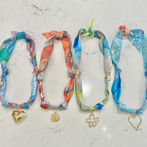 May include: Four patterned silk scarf necklaces with gold pendants. The scarves display various designs in blue, orange, green, and white. Each necklace has a different gold pendant: a heart, a coin, a flower, and a heart outline.